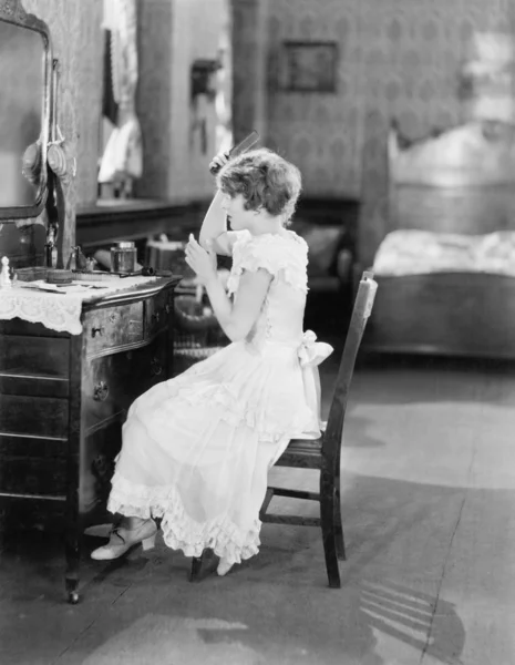 Jovem mulher sentada na frente de sua mesa de vaidade e arrumando seu cabelo — Fotografia de Stock