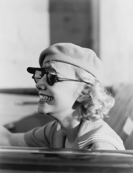 Ung kvinna med en basker och solglasögon i en bil, på språng — Stockfoto