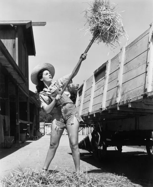 Giovane donna che indossa jeans tagliati e lavora alla fattoria lanciando fieno in un carro — Foto Stock