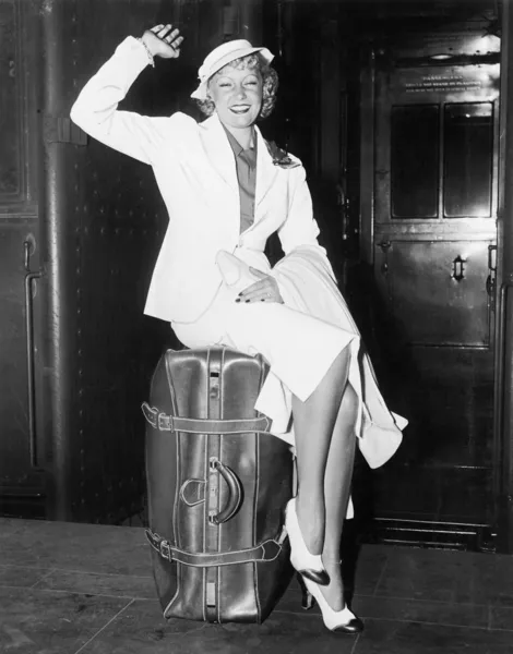 En klar att gå ung kvinna sitter på hennes resväska och vinkar — Stockfoto