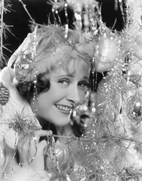 Jovem mulher olhando através dos ramos de uma árvore de Natal com ornamentos — Fotografia de Stock