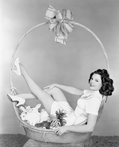 Jovem mulher, parecendo um presente, sentada em cesta de presente com coelhos — Fotografia de Stock