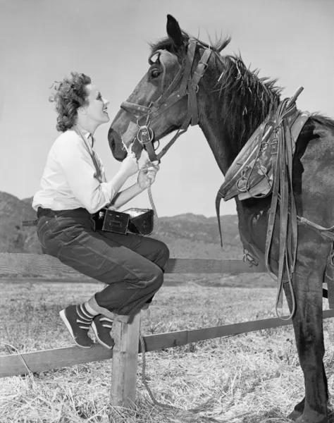 Mujer joven sentada en una valla y alimentando a su caballo — Foto de Stock