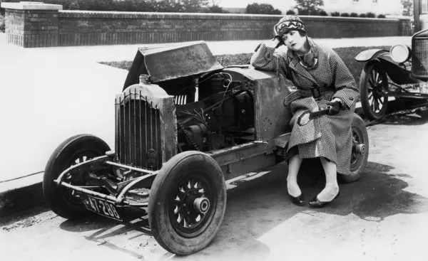 Νεαρή γυναίκα που κάθεται δίπλα στο μικρό αυτοκίνητο κοιτάζοντας τον κινητήρα στο δυσπιστώ — Φωτογραφία Αρχείου