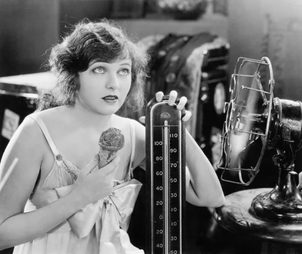 Mujer joven sentada junto a un ventilador y un termómetro que luce caliente y come un helado —  Fotos de Stock
