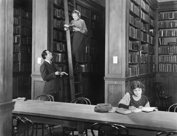 Чоловік розмовляє з жінкою, стоячи на драбині в бібліотеці — стокове фото