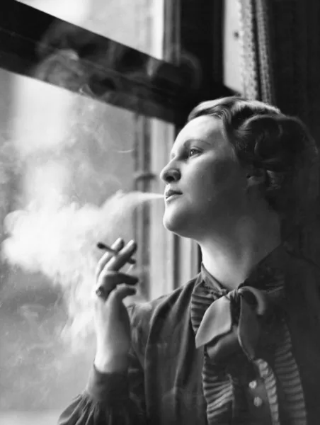 Νεαρή γυναίκα που κάθεται δίπλα σε παράθυρο, κάπνισμα — Φωτογραφία Αρχείου
