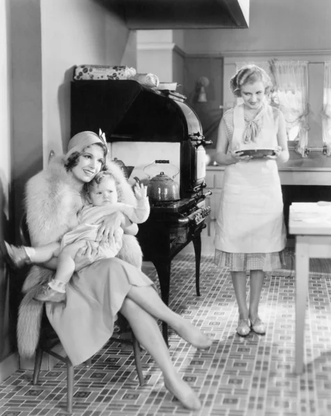 Elegáns nő ül a konyhában gyermekével, míg egy barátja a torta hozza — Stock Fotó
