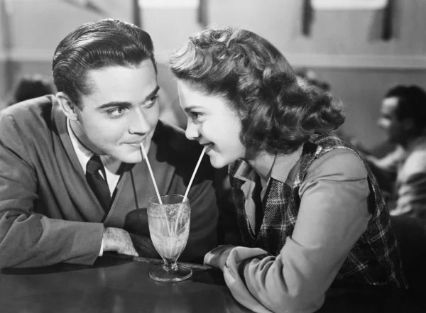 Пара в ресторане смотрит друг на друга и делится молочным коктейлем с двумя соломинками Стоковое Фото