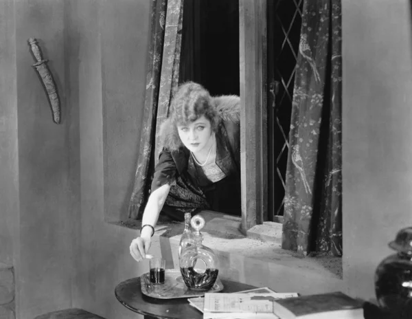 Портрет молодой женщины, проникающей в окно и наливающей яд в стакан Стоковая Картинка