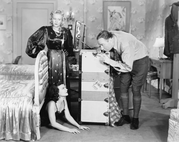 Young woman hiding under a bed and a couple standing near her Stock Picture