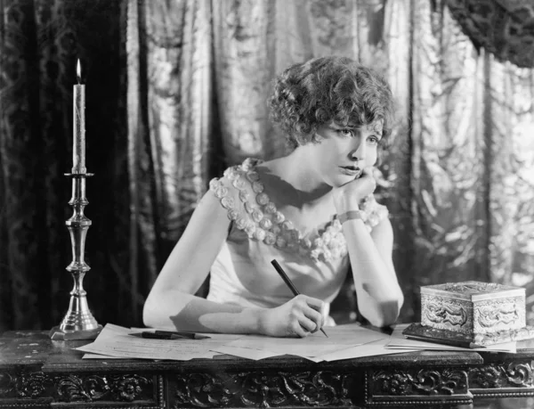 Mujer joven sentada en un escritorio con un bolígrafo en la mano, mirando triste mientras escribe una carta Imagen de archivo