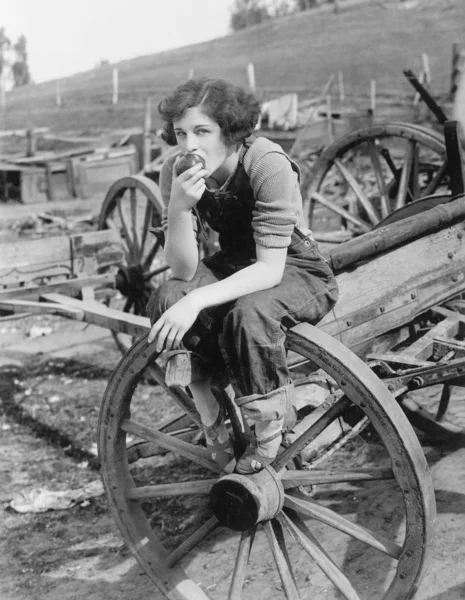 Jeune femme en jeans de travail assis sur la roue manger une pomme Images De Stock Libres De Droits