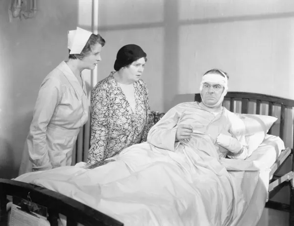 A wife next to her husband in a hospital bed with a nurse attending Stock Photo