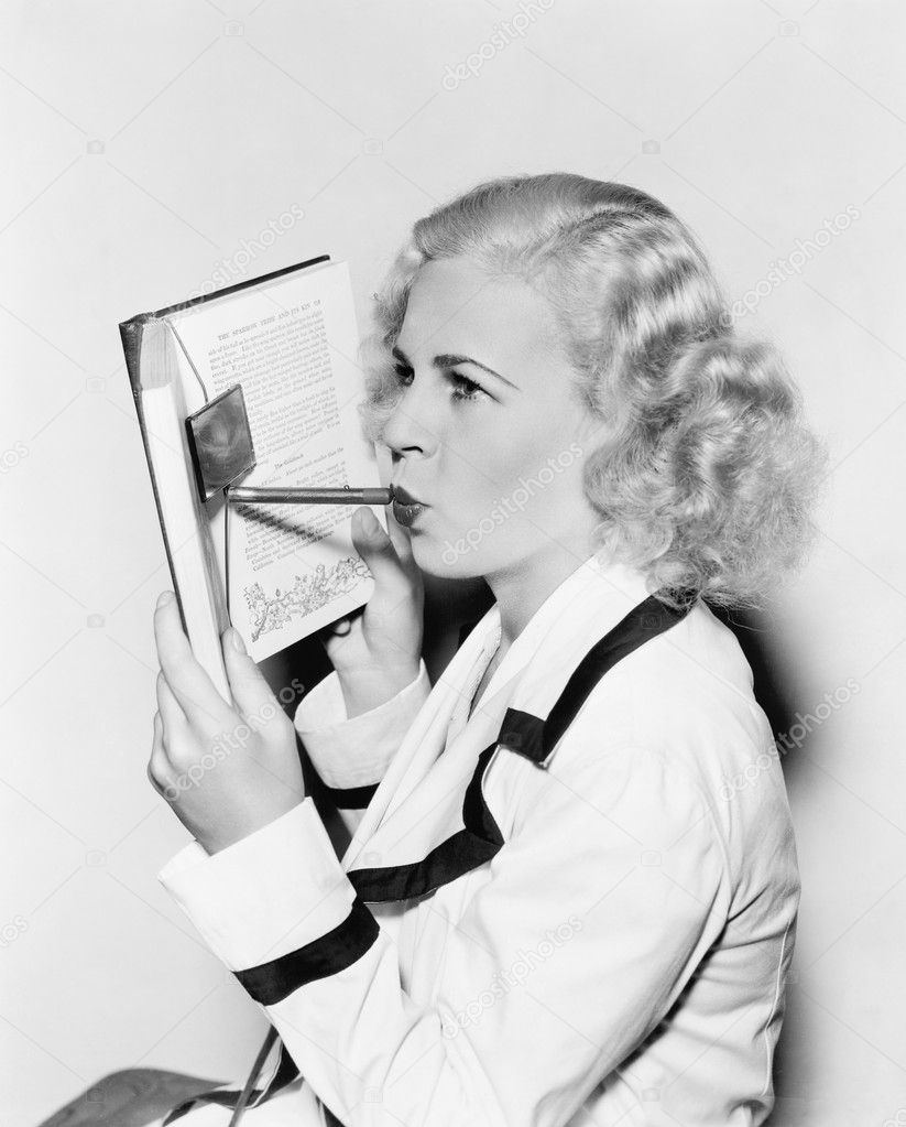 Young woman hiding her face behind a book applying lipstick
