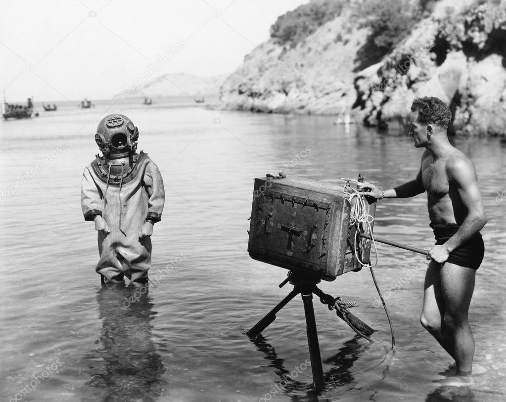 Profile of a young man holding a camera with a scuba diver standing in front of him on the beach