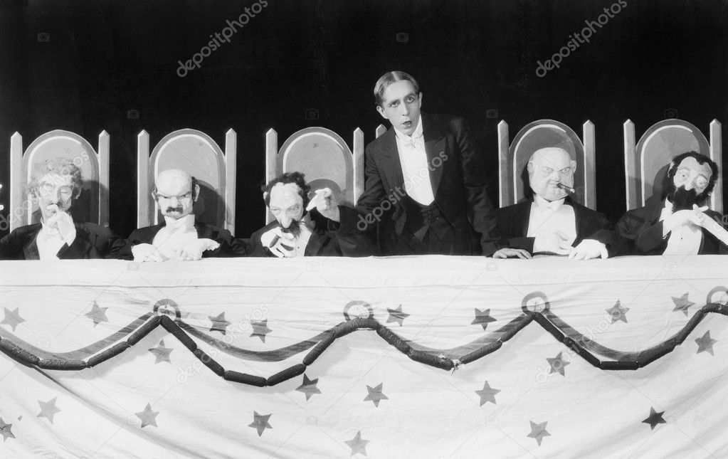 Man giving a speech with five character puppets sitting at the table