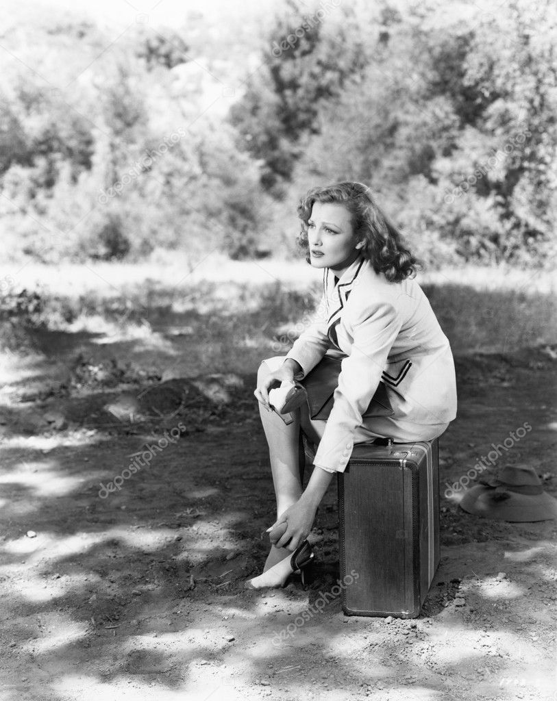 Young woman sitting on the side of the road on her suitcase, rubbing her aching feet