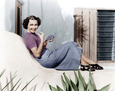 Young woman sitting on a terrace smiling and eating grapes clipart