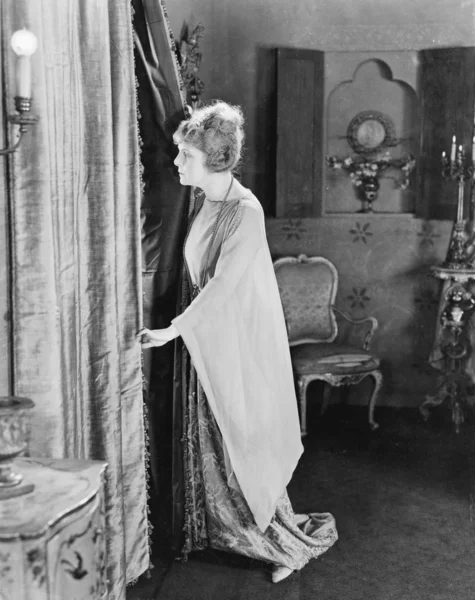 Mujer joven mirando desde detrás de sus cortinas — Foto de Stock