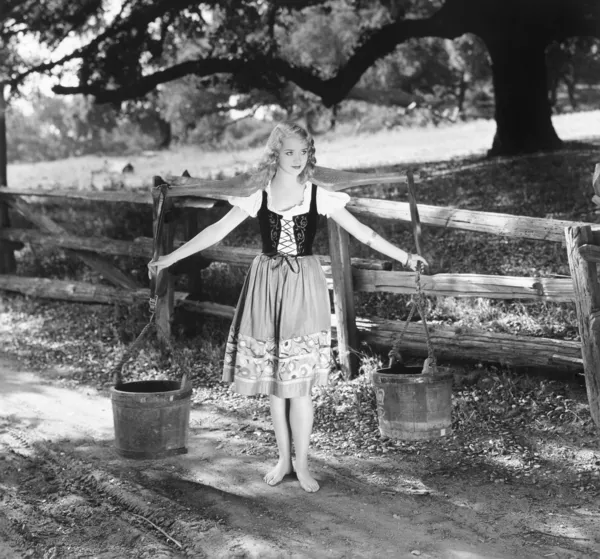 Mulher descalça em um vestido de corpete amarrado carregando baldes de água em postes de ombro — Fotografia de Stock