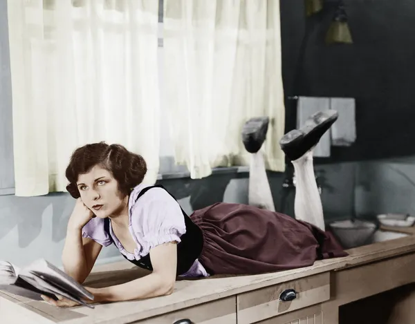 Mladá žena leží na kuchyňské lince drží knihu a myšlení — Stock fotografie