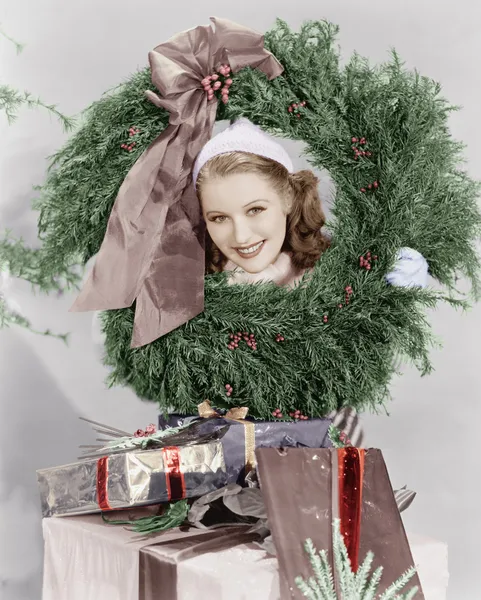 Young woman looking through a wreath with presents in front of her — Stock Photo, Image
