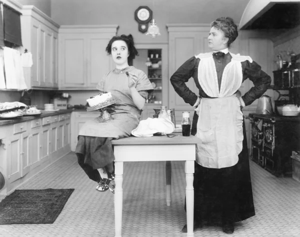 Haushälterin in der Küche starrt eine junge Frau an, die einen Kuchen isst Stockbild