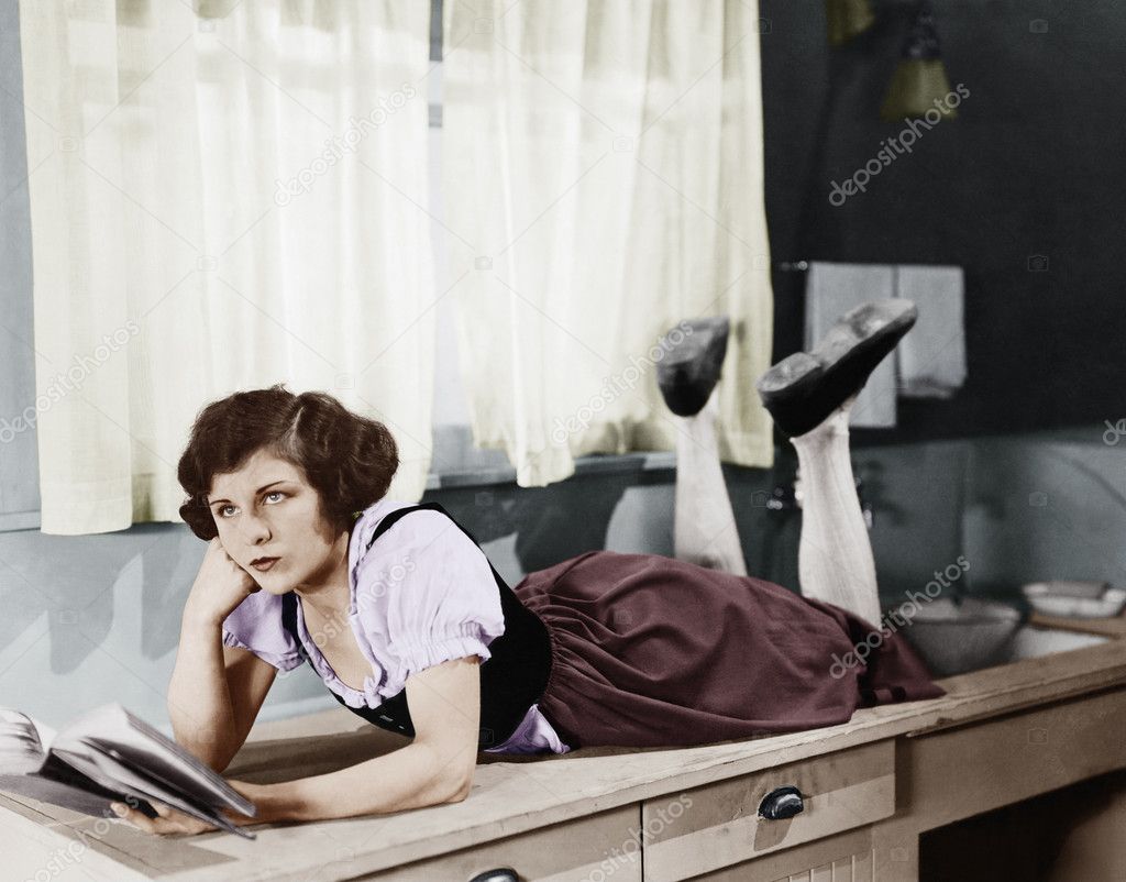 Young woman lying on a kitchen counter holding a book and thinking