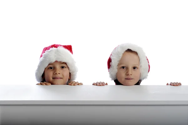 Zwei süße kleine Jungen mit Weihnachtsmütze schauen hinter einem Tisch hervor — Stockfoto