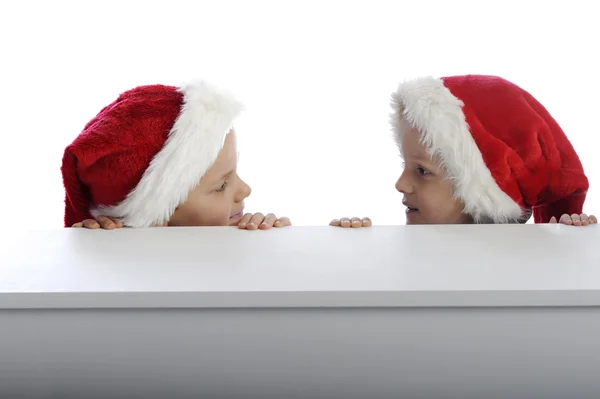 Two cute little boys with xmas hat looking from behind a table — Stock Photo, Image